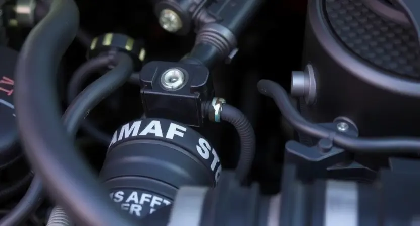 A close-up, detailed image of a car's engine bay, specifically focusing on the Mass Airflow Sensor (MAF) and its wiring. The MAF sensor should be clearly visible, along with the surrounding hoses and electrical connectors. The image should convey a sense of technical inspection and diagnostics. Focus on the sensor's physical appearance and the condition of the wiring.